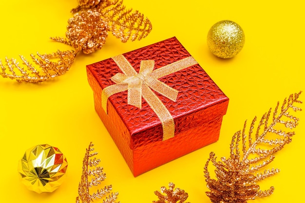 Red gift box, golden balls on the christmas tree and shiny twigs with cones on a yellow background