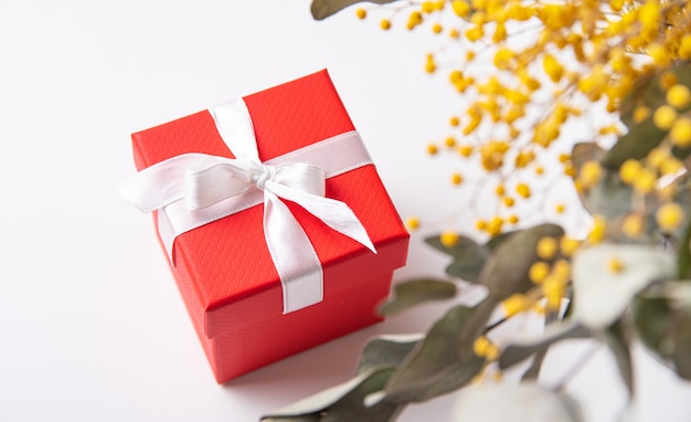 Красная подарочная коробка с белой лентой на белом столе под весенними цветами мимозы. Вид сверху