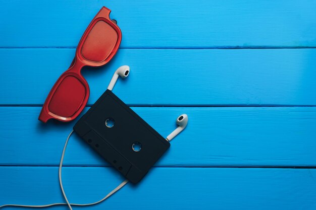 red glasses classic magnetic cassette tape and white headphones on a blue wooden table background