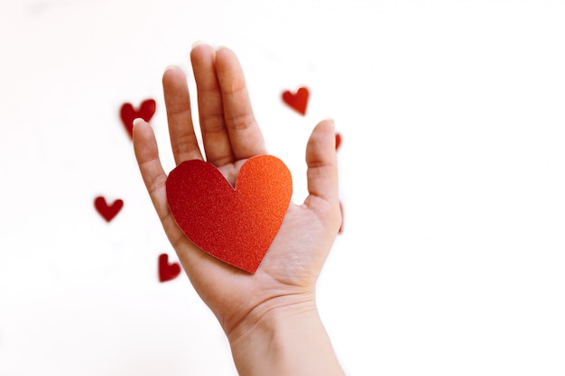 Red glitter heart shaped cardboard on the hand and small hearts on white surface. Concept of love and valentines day.