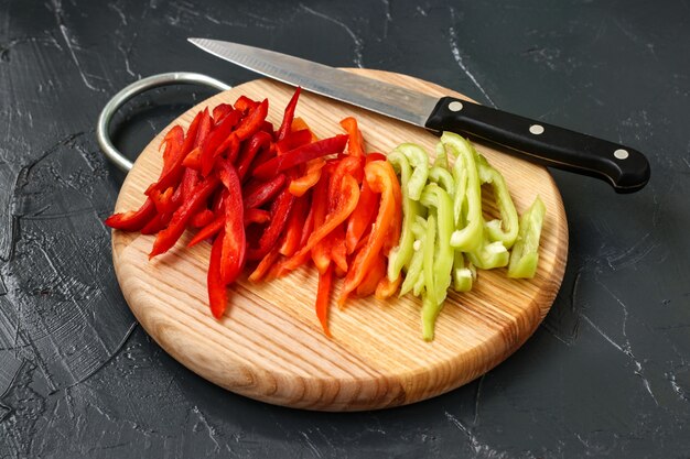 Red and green sweet pepper sliced in strips placed on a wooden board on a dark surface, Top view