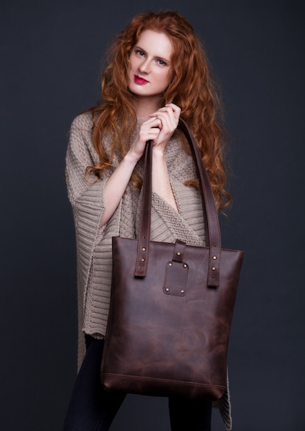 Red hair fashion model holding large dark leather bag on dark background. Girl wearing jumper.