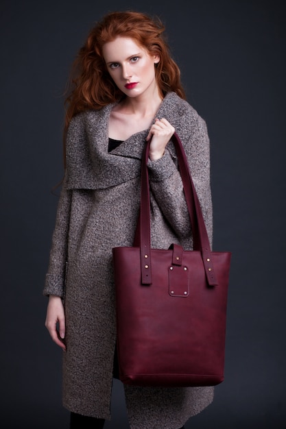 Red hair fashion model holding large dark red leather bag on dark background. Girl wearing long jumper.