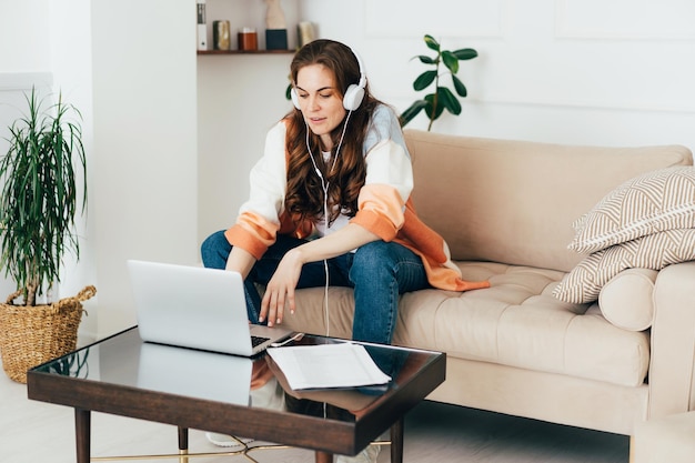 Red-haired attractive young lady works online in zoom using a laptop and headphones