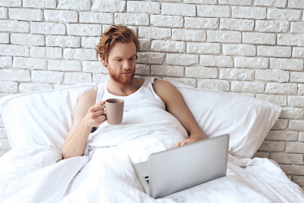 Red haired awaking man lies in bed with laptop.