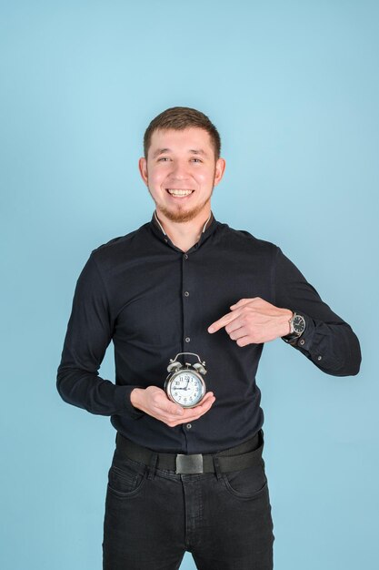 A red-haired bearded man holds an alarm clock in his hands and points at it with his finger standing on a blue background, vertical photo