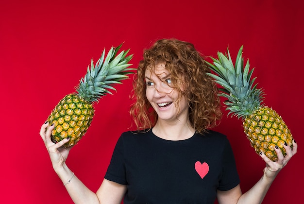 Bella dai capelli rossi in una maglietta nera con una stampa di cuore tiene un ananas tra le mani su uno spazio rosso