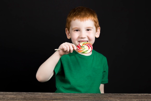 美しい顔の赤い髪の少年は、棒で甘いマルチカラーのキャンディーを食べます、砂糖で作られたロリポップを持つ少年、彼の歯に虫歯を持つ少年