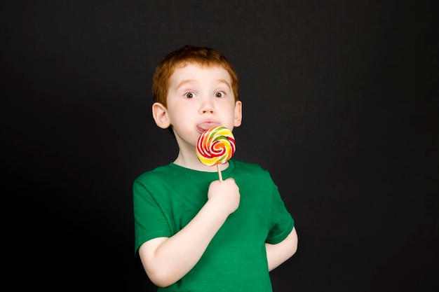 美しい顔をした赤毛の少年は、甘いマルチカラーのキャンディーを棒で食べます。砂糖でできたロリポップを手に持った少年です。