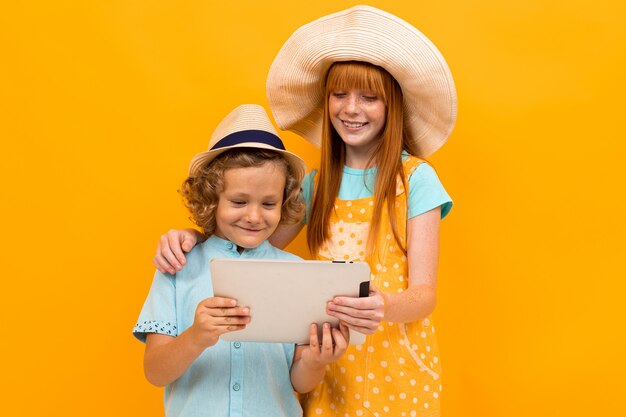 Red-haired brother and sister in summer hats book tickets on a tablet on a yellow background.