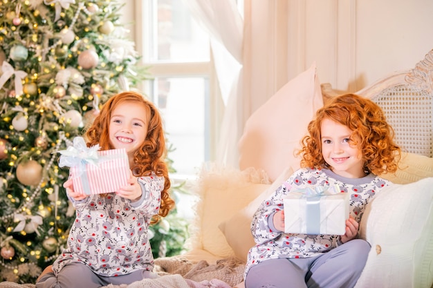 Red-haired children in pajamas sitting on the bed with gifts in their hands against the Christmas tree.