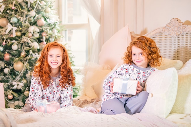 Red-haired children in pajamas sitting on the bed with gifts in their hands of the Christmas tree.