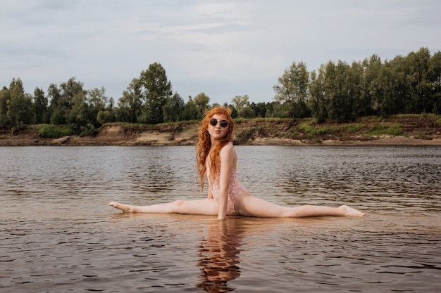 A red-haired girl in a bathing suit is sitting on a splits in shallow water on a summer day.