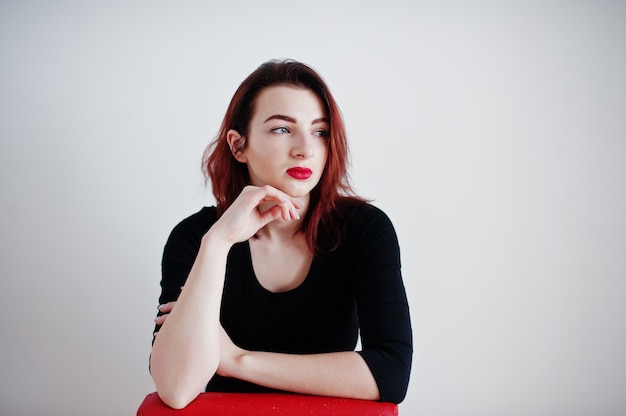 Red haired girl on black dress tunic sitting on red chair against white wall at empty room.