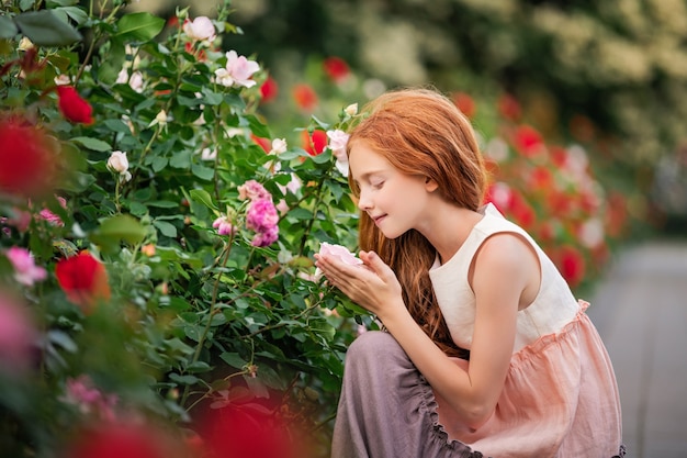 Red-haired girl child sniffing a rose in blooming garden in summer