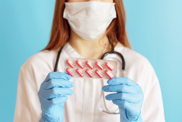 a red-haired girl doctor in a protective face mask and medical gloves with a stethoscope shows capsules tablets with antiviral medication, a concept of protection against colds, viruses and bacteria