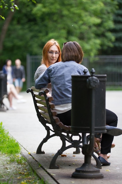 안경을 쓴 빨간 머리 소녀는 여자 친구 옆 공원 벤치에 앉아 있습니다.