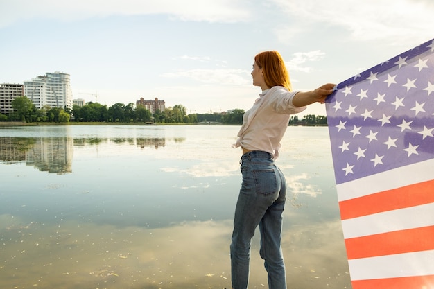Red haired girl holding USA national flag in her hands. Positive young woman celebrating United States independence day.