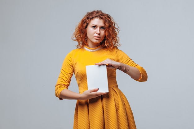 Red-haired girl holding a white book on a light gray background.