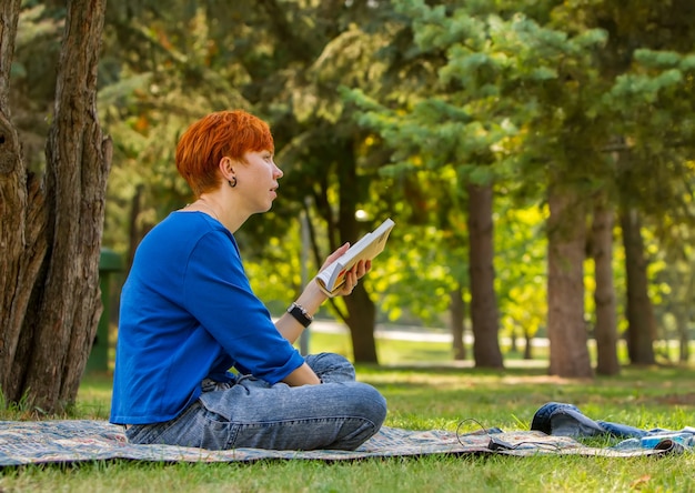 Red-haired girl reads a book in a clearing in the park and looks thoughtfully into the distance.