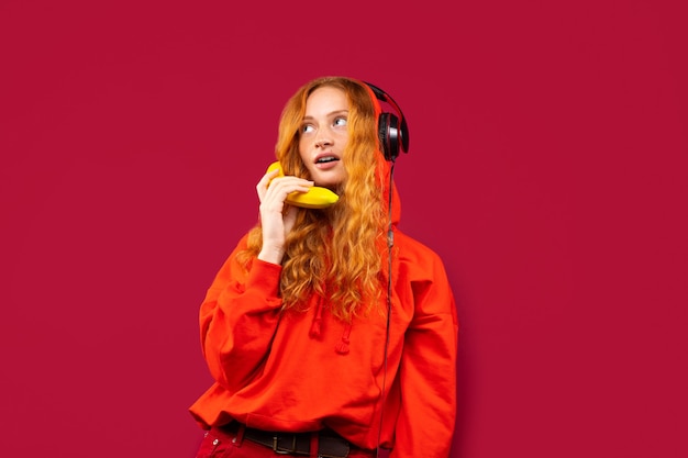 Una ragazza dai capelli rossi con una camicia rossa e grandi cuffie, ascolta musica e posa con una banana. foto su un muro rosso, il concetto di ricreazione e passatempo.