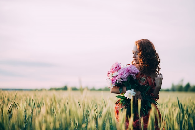 Red-haired girl in red dress with bouquet of peonies flowers in summer wheat field at sunset. 