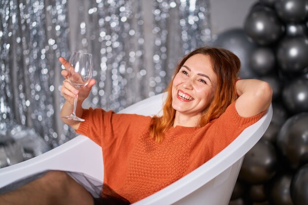 Red-haired girl rejoices sitting dressed in the bath