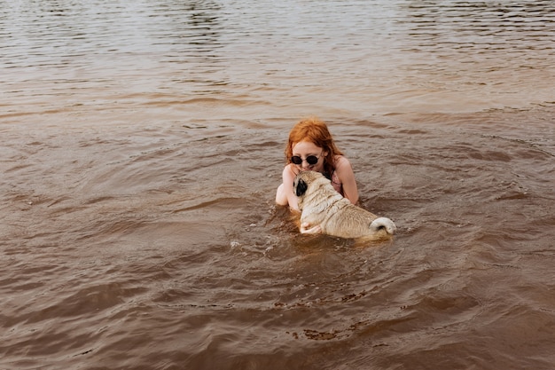 Red-haired girl teaches her pug to swim in the river.