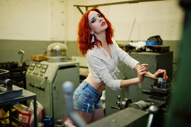 Red haired girl wear on short denim shorts and white blouse posed at industrial machine at the factory