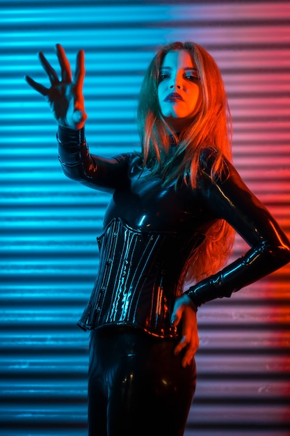 Red-haired girl wearing latex, a black top and tight pants, on a metallic background illuminated with red and blue LEDs, making the stop gesture