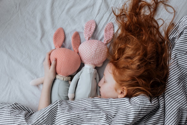 Red-haired girl with dolls on the bed.