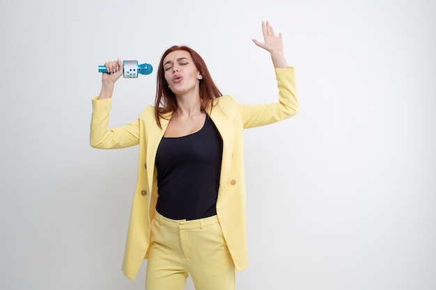 Red-haired girl in a yellow suit sings into a microphone on a white background