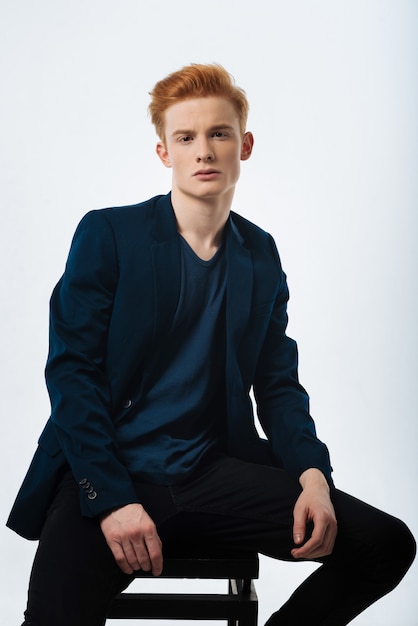 Red-haired. Handsome unsmiling red-haired young man wearing a black jacket and staring and sitting on the chair