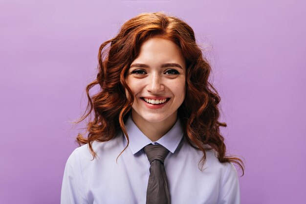 Red haired lady in shirt and tie poses on purple wall