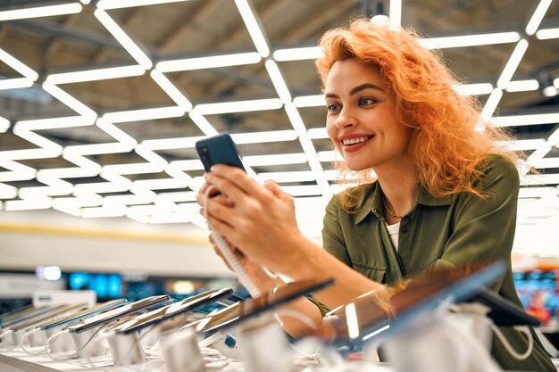 Red-haired sweet woman chooses new smartphone in store of\
household appliances, electronics and gadgets. buying new stylish\
smartphone.