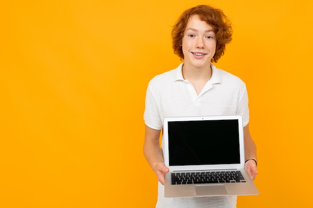 Red-haired teenager in a white t-shirt holds a laptop on a yellow wall