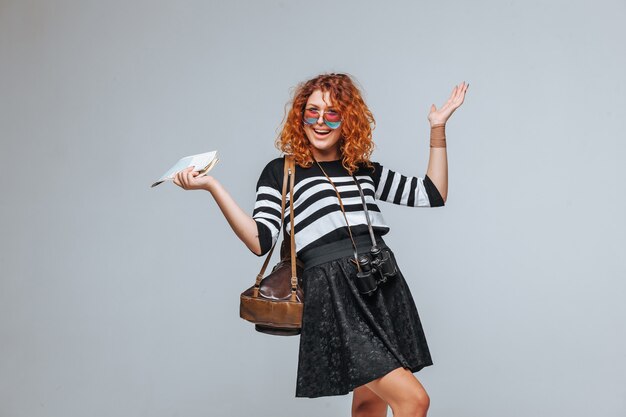 Red-haired tourist girl with a map, binoculars and backpack on a gray light background