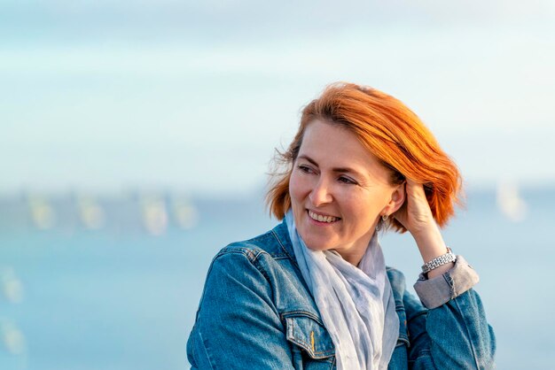 Red haired woman in blue dress and jeans jacket resting on the seashore at sunset