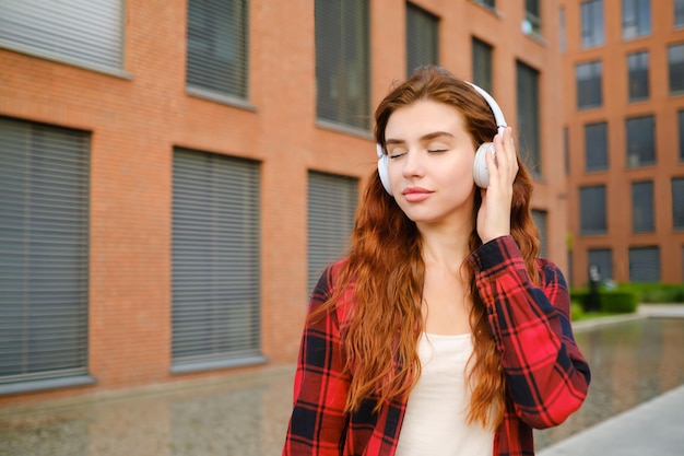 赤い石の建物の近くに立って音楽を楽しむヘッドフォンの赤髪の女性