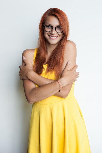 Red haired woman with glasses is embracing herself wearing a yellow dress on a white  wall smiling 