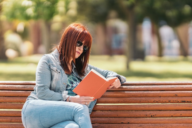 Red-haired woman with sunglasses reading a book sitting sideways on a park bench on a sunny day