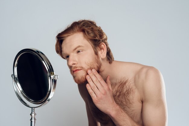 Red haired young man strokes beard, looking into mirror.