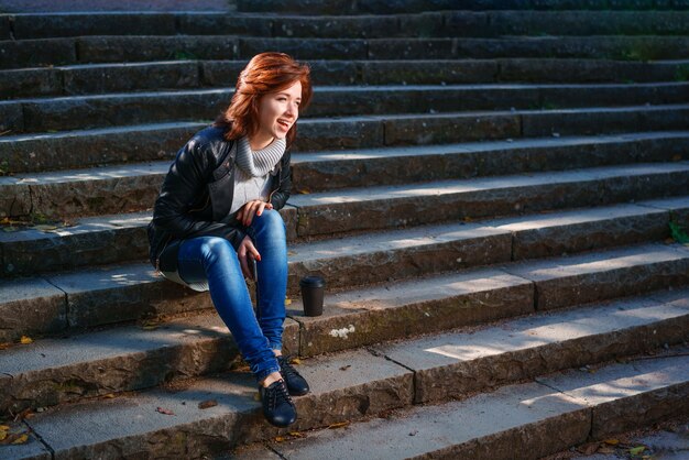 Una giovane donna dai capelli rossi in giacca di pelle e jeans si siede sui gradini con una tazza di caffè e si sveglia.