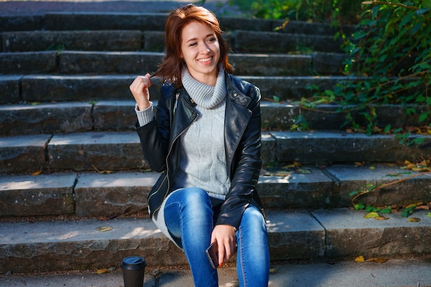 A red-haired young woman in a leather jacket and jeans sits on the steps with a cup of coffee