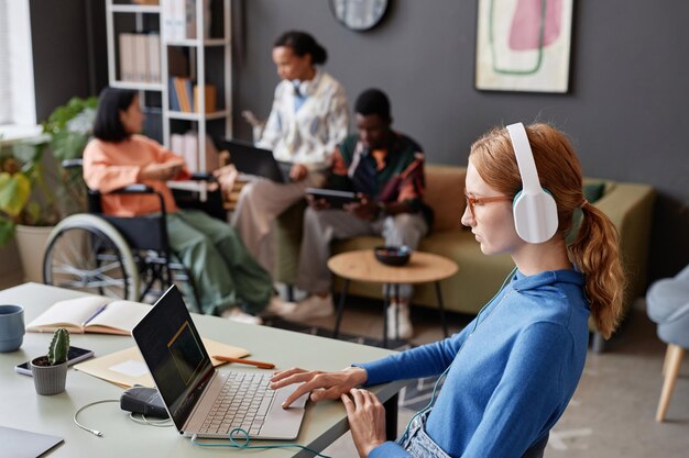 Photo red haired young woman wearing headphones in office working with laptop