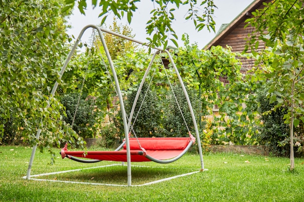 Red hammock swing in metal frame with nobody on green lawn in backyard near log house cottage. Rest relax relaxation alone on hammock swing in Summer garden.