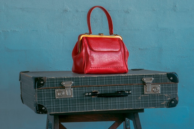 A red handbag stands on a vintage travel suitcase against a blue concrete wall