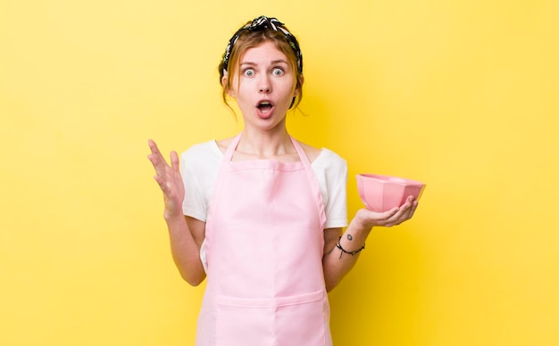 Red head pretty woman amazed shocked and astonished with an unbelievable surprise housewife holding an empty bowl