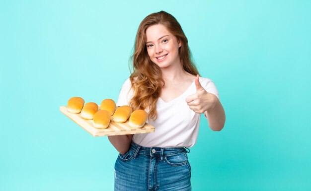 Red head pretty woman feeling proud,smiling positively with thumbs up and holding a bread buns troy