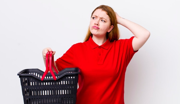 Red head pretty woman feeling puzzled and confused scratching head empty shopping basket concept
